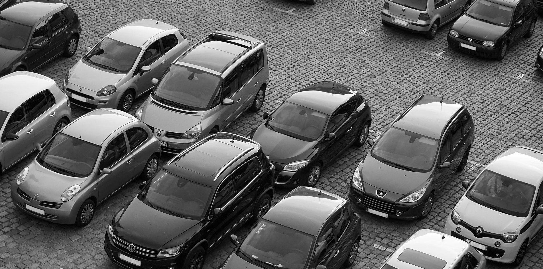 Vista dall'alto in bianco e nero di auto Volkswagen, Peugeot e Renault ferme su un piazzale lastricato con cubetti