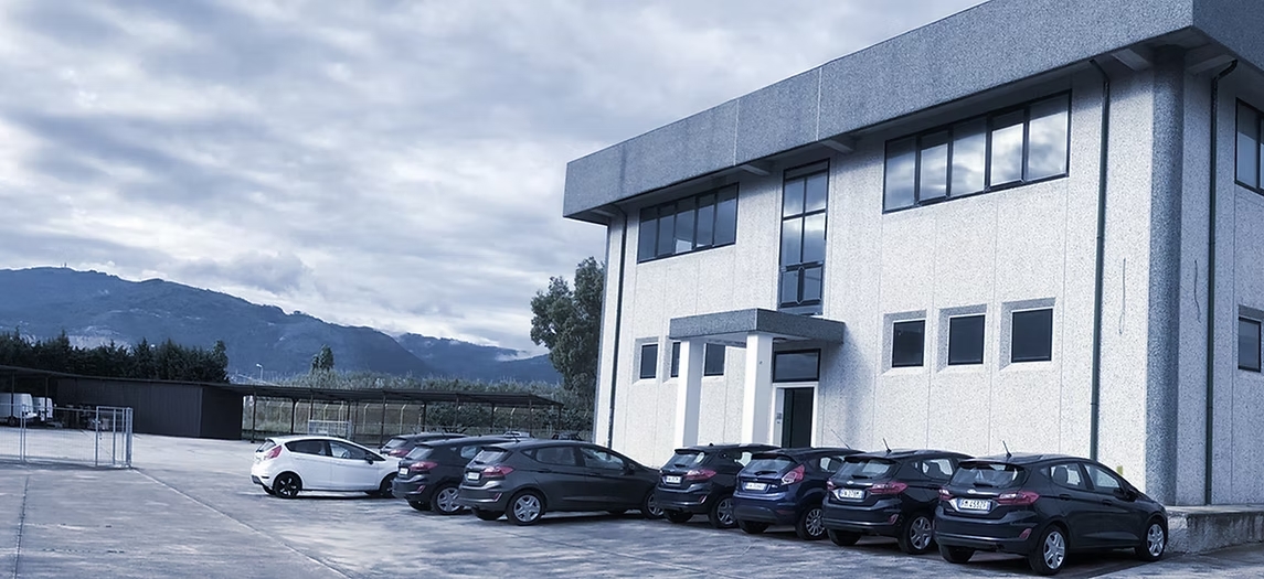 Utilitarie scure e una bianca davanti alla sede di Security Parking, con tettoie e montagne sullo sfondo