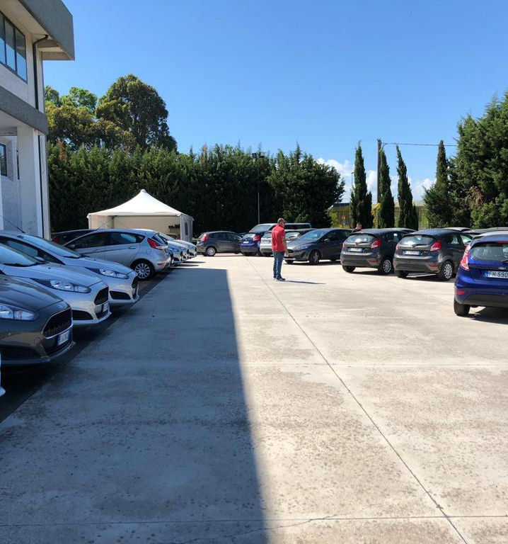 Piazzale di cemento di Security Parking con file di utilitarie, gazebo bianco, siepe alta, cipressi e un uomo in maglia rossa
